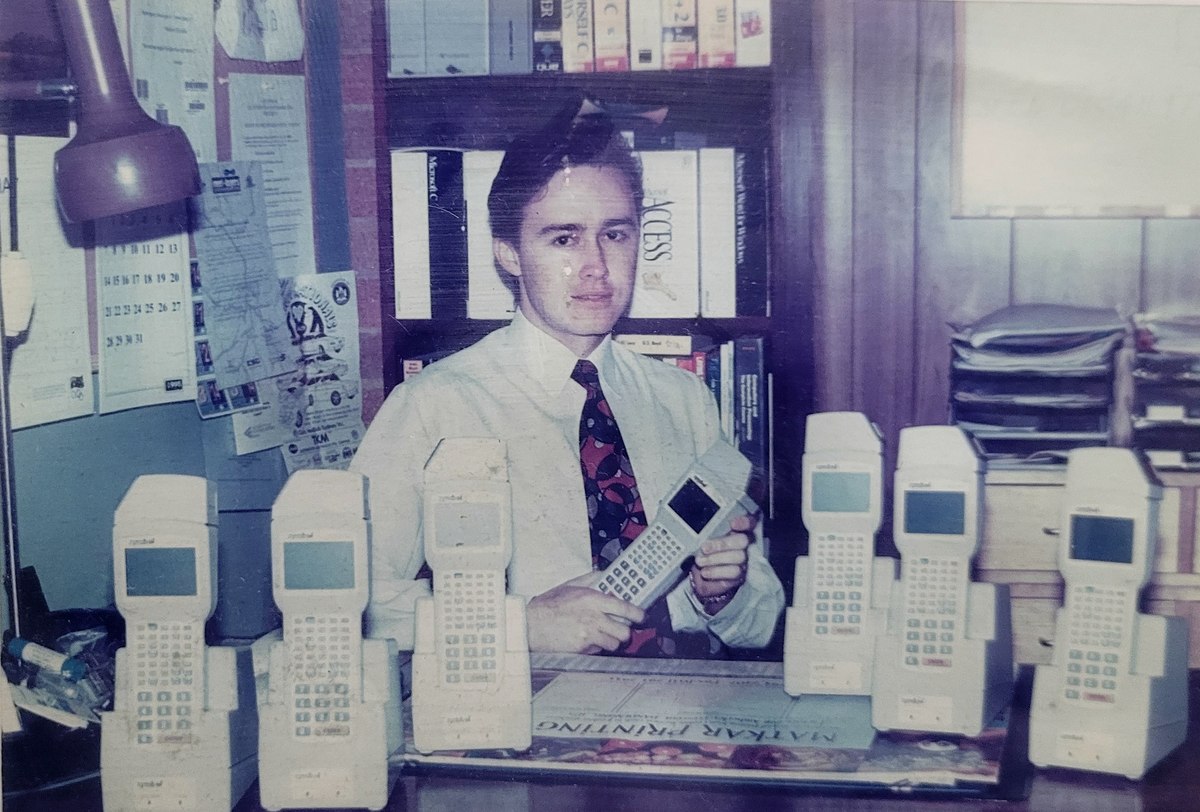 Andrei Spiegel in 1995 in his garage home office, behind seven Symbol
                  PDT3100 terminals on a desk, holding an eighth