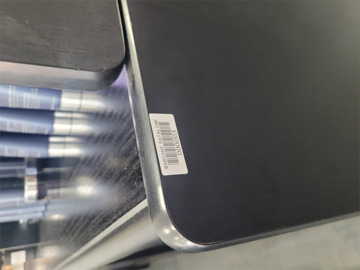 An asset label reading BARCODE DATALINK and the number 000003 on the edge of a dark display cabinet, with shelving and stock behind it.