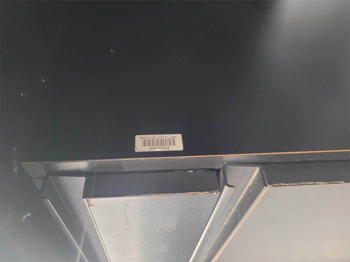 An asset label reading BARCODE DATALINK and the number 009050 on the painted underside of a dark desk.