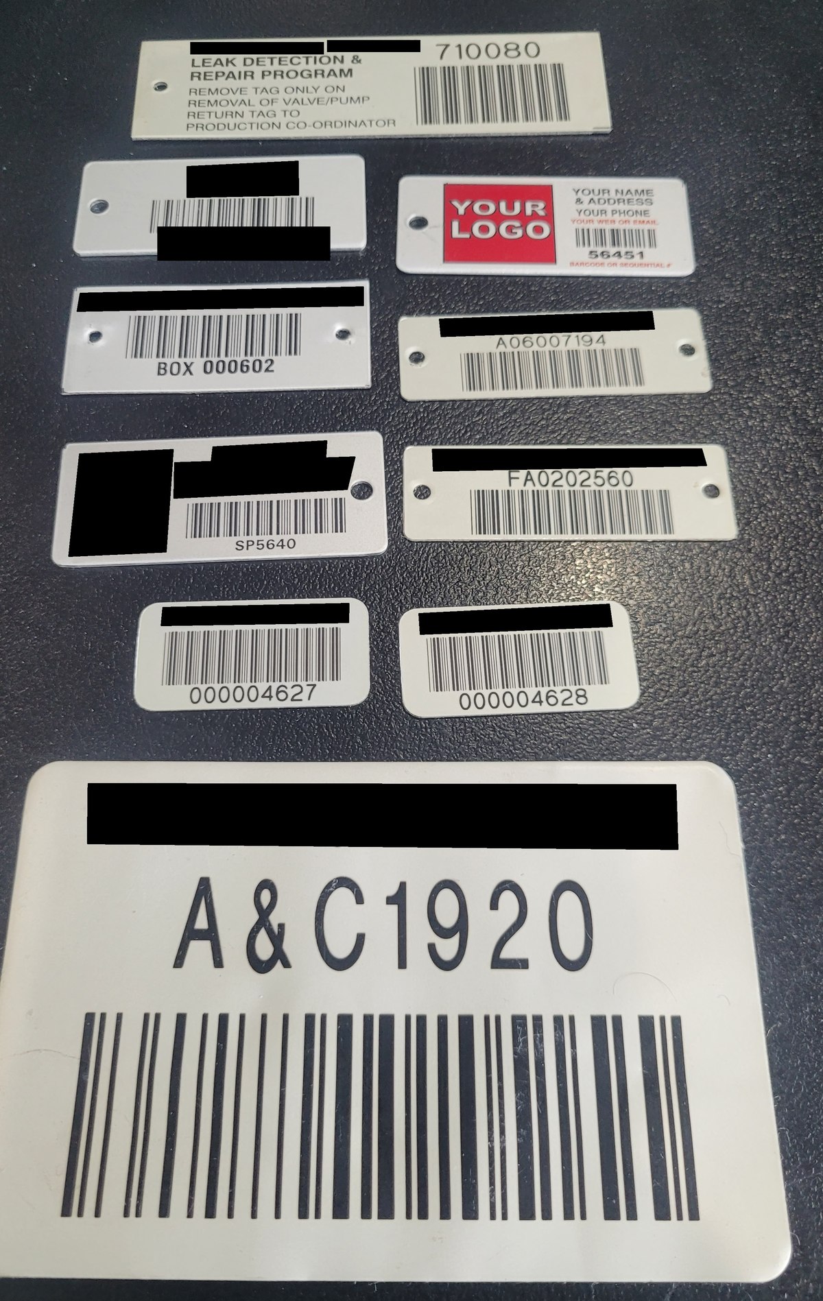 Nine aluminium asset labels and tags laid out on a bench in various sizes, some
                  with screw holes and some without, showing barcodes, sequential numbers and
                  instruction text. Company names have been blacked out.