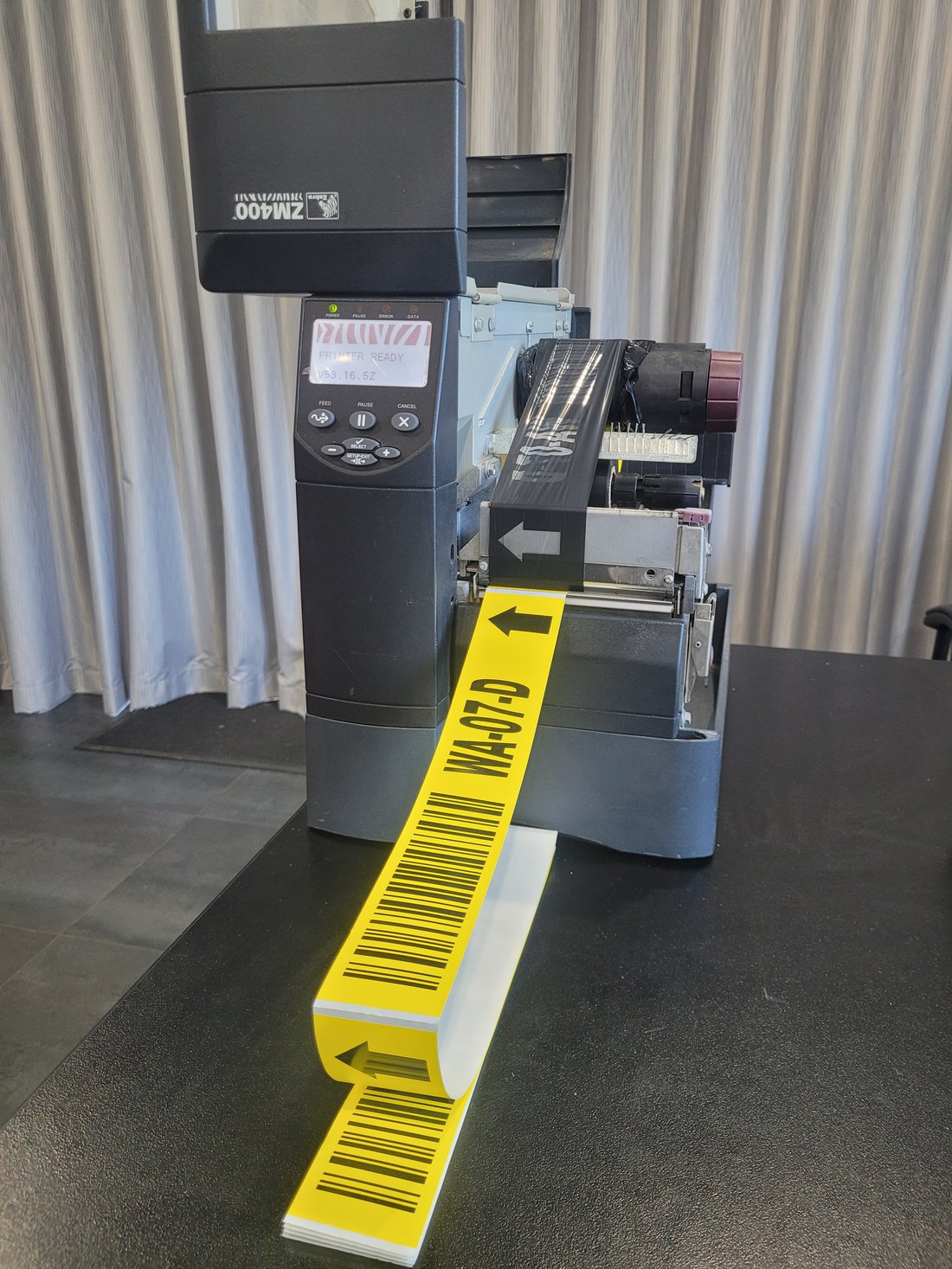 A Zebra ZM400 printing yellow bin location labels which fold flat onto the bench, each reading WA-07-D with a barcode and a left-pointing arrow.