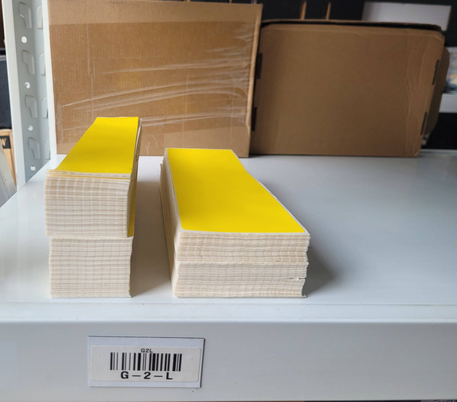 Two stacks of yellow fanfold labels folded flat on a white warehouse shelf, with a barcoded bin label reading G-2-L on the shelf edge.