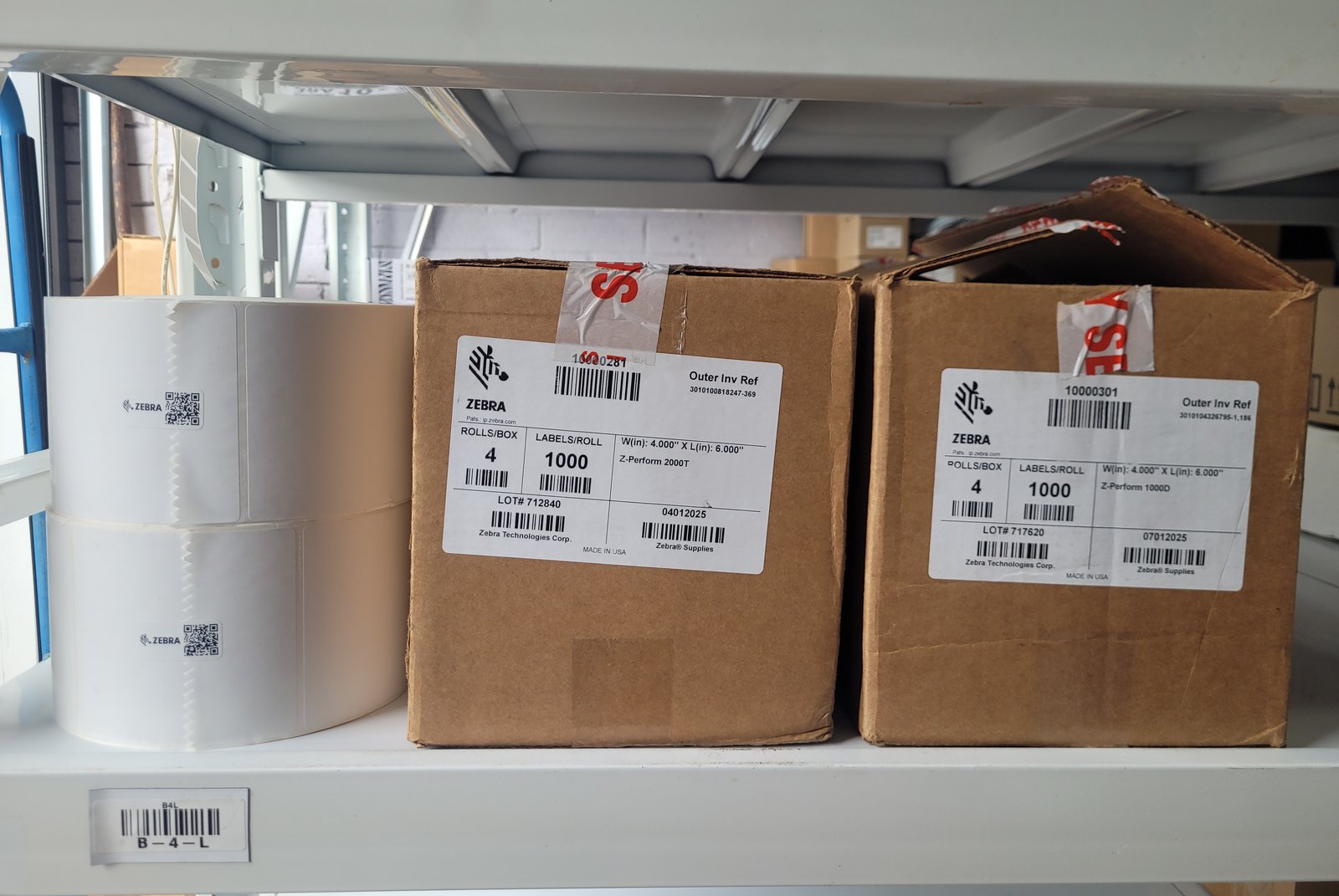 Two sealed Zebra label cartons on a white warehouse shelf beside an open roll of white labels, with a barcoded bin label reading B-4-L on the shelf edge. Both carton labels read ROLLS/BOX 4 and LABELS/ROLL 1000.