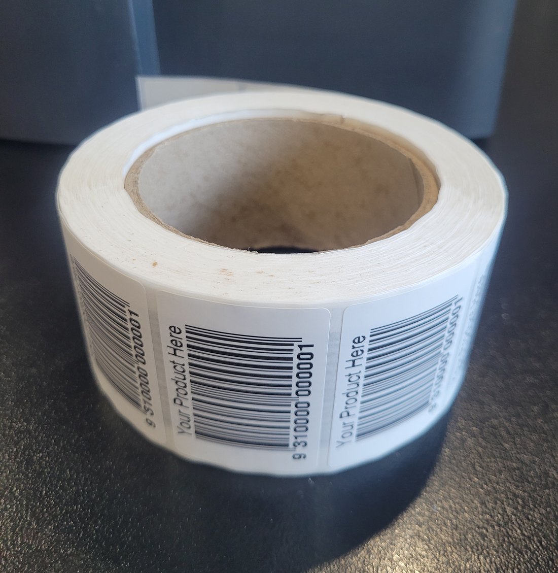 A finished roll of printed labels on a cardboard core, wound and sitting on a bench, each label reading Your Product Here above a barcode.