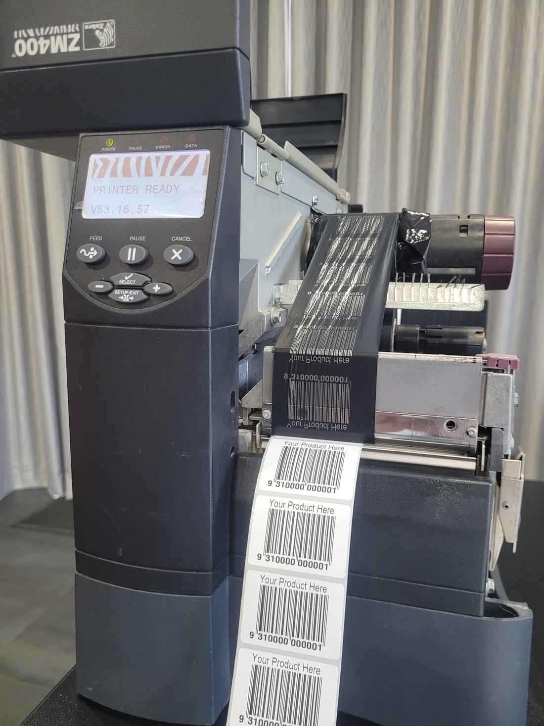 A Zebra ZM400 industrial printer with its cover open, display reading PRINTER READY. The used ribbon carries a mirrored image of every label printed, and a strip of finished labels comes out of the front.