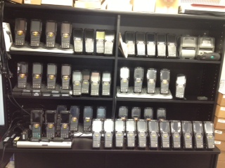 Shelving racked with roughly forty Symbol MC9000 series handheld scanners
                  standing in charging cradles, three shelves deep, with a printer at one end.