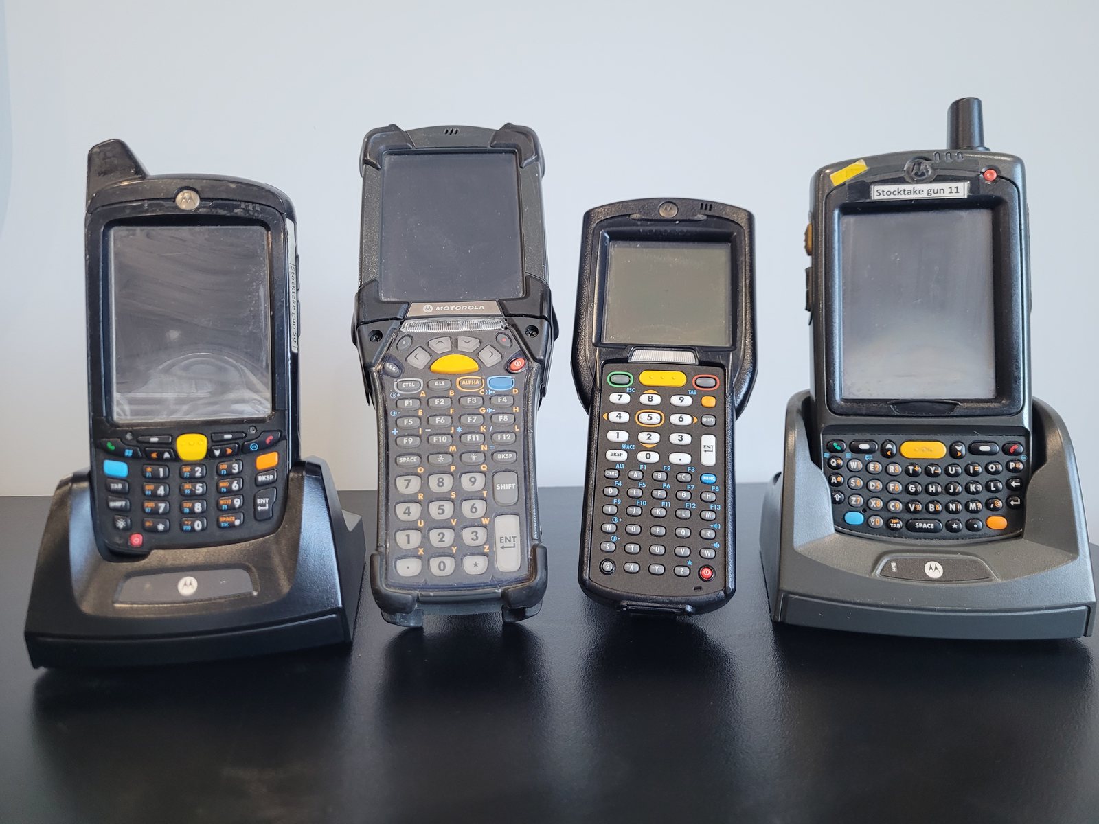 Four Motorola Windows Mobile handheld computers side by side on a black
                    bench: an MC65 and an MC75 sitting in charging cradles, and an MC9200 and
                    MC3100 standing between them