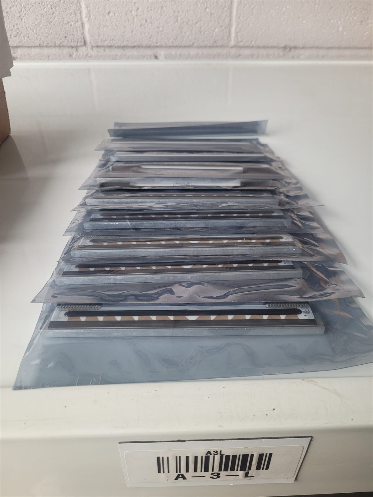 A row of about ten bare thermal printheads, each sealed in a clear anti-static bag, laid out on a white shelf beside a barcoded bin label reading A-3-L.