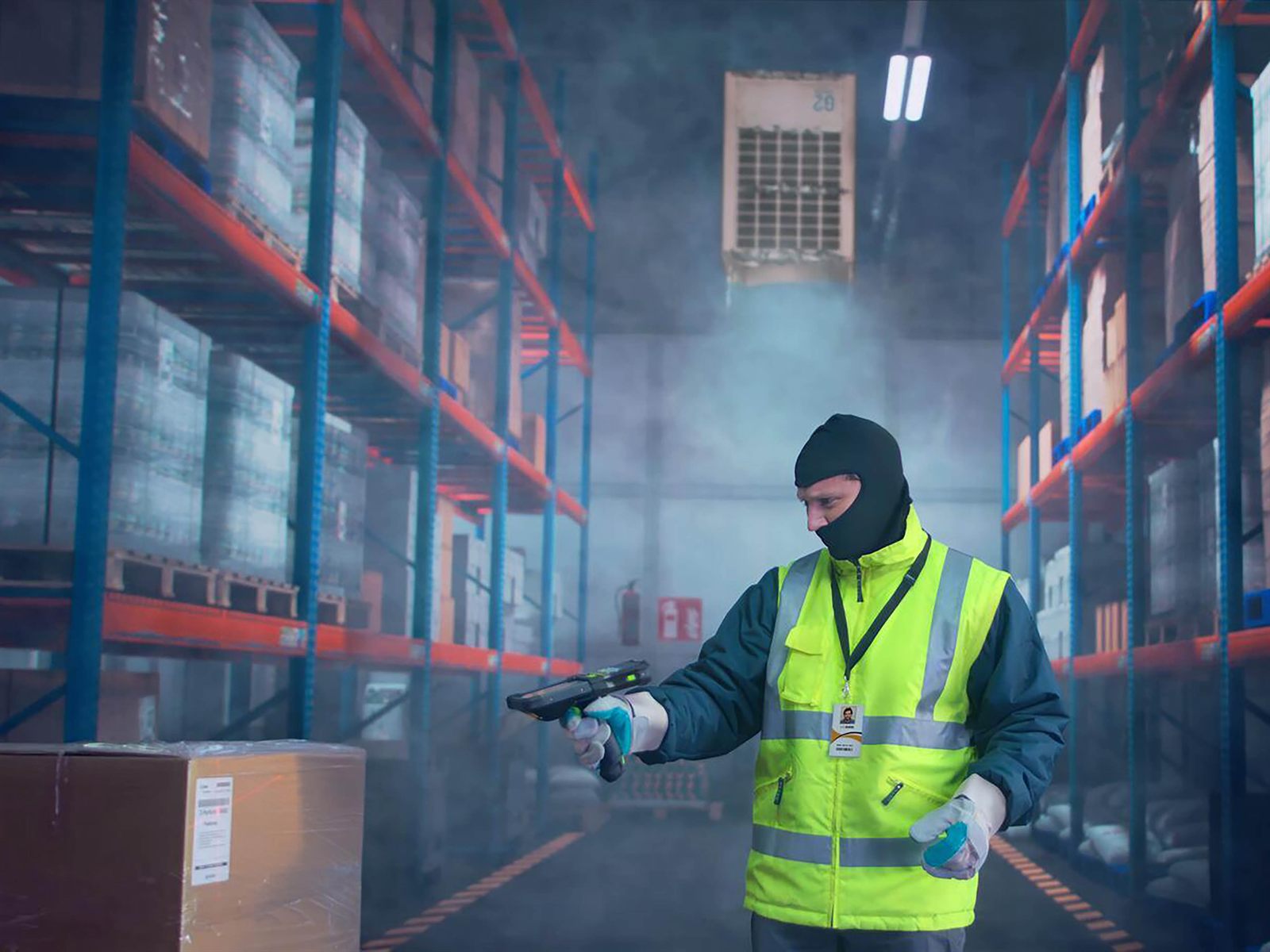 An operator in a freezer jacket, balaclava and thermal gloves standing in a
                    cold store aisle with fog hanging under the evaporator, scanning with a
                    gun-grip Zebra MC9400.