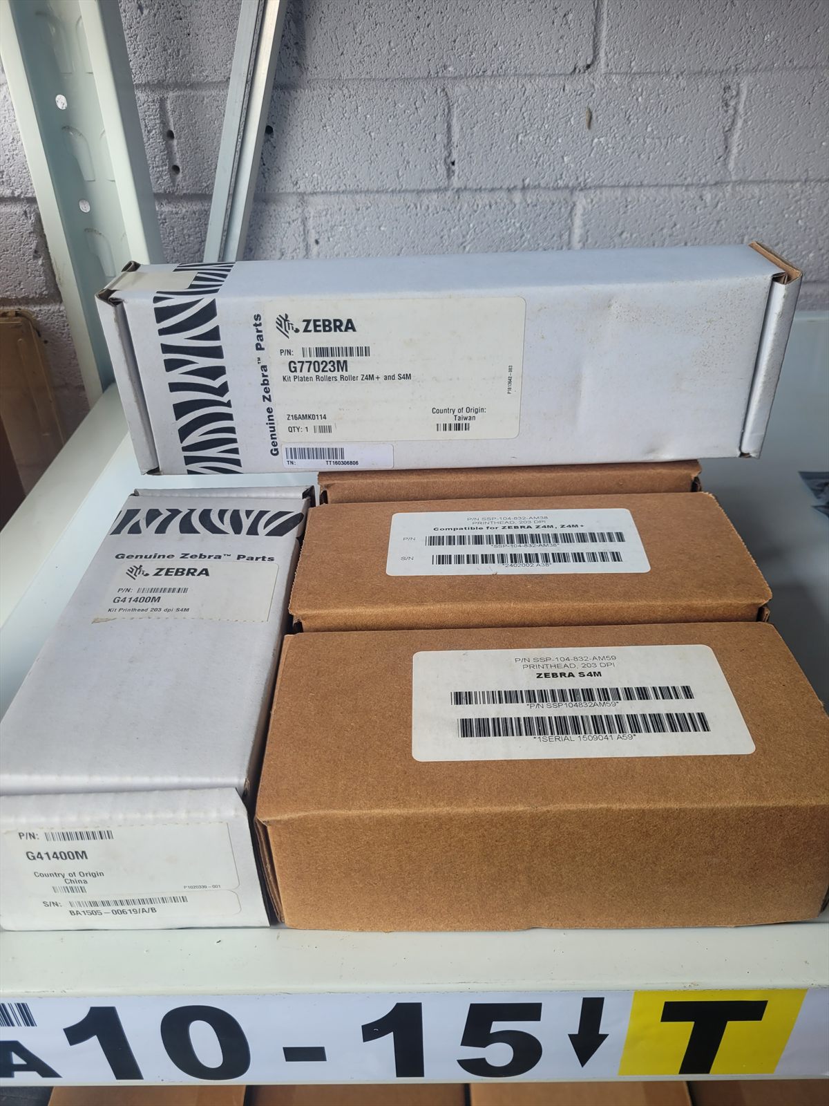 Close view of four boxes on the shelf. Two are white Genuine Zebra Parts cartons labelled G77023M and G41400M; two are plain brown boxes with printed part number labels.