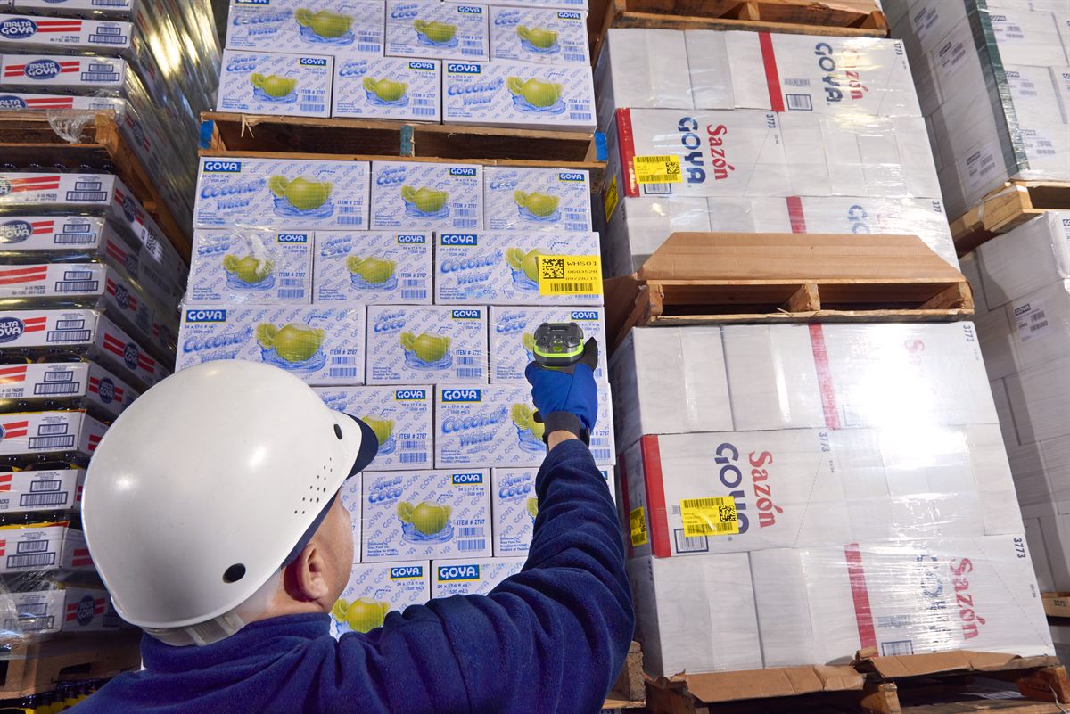 A warehouse worker in a hard hat reaching up with a rugged green Zebra scanner to read a yellow 2D location label on a stack of pallets.