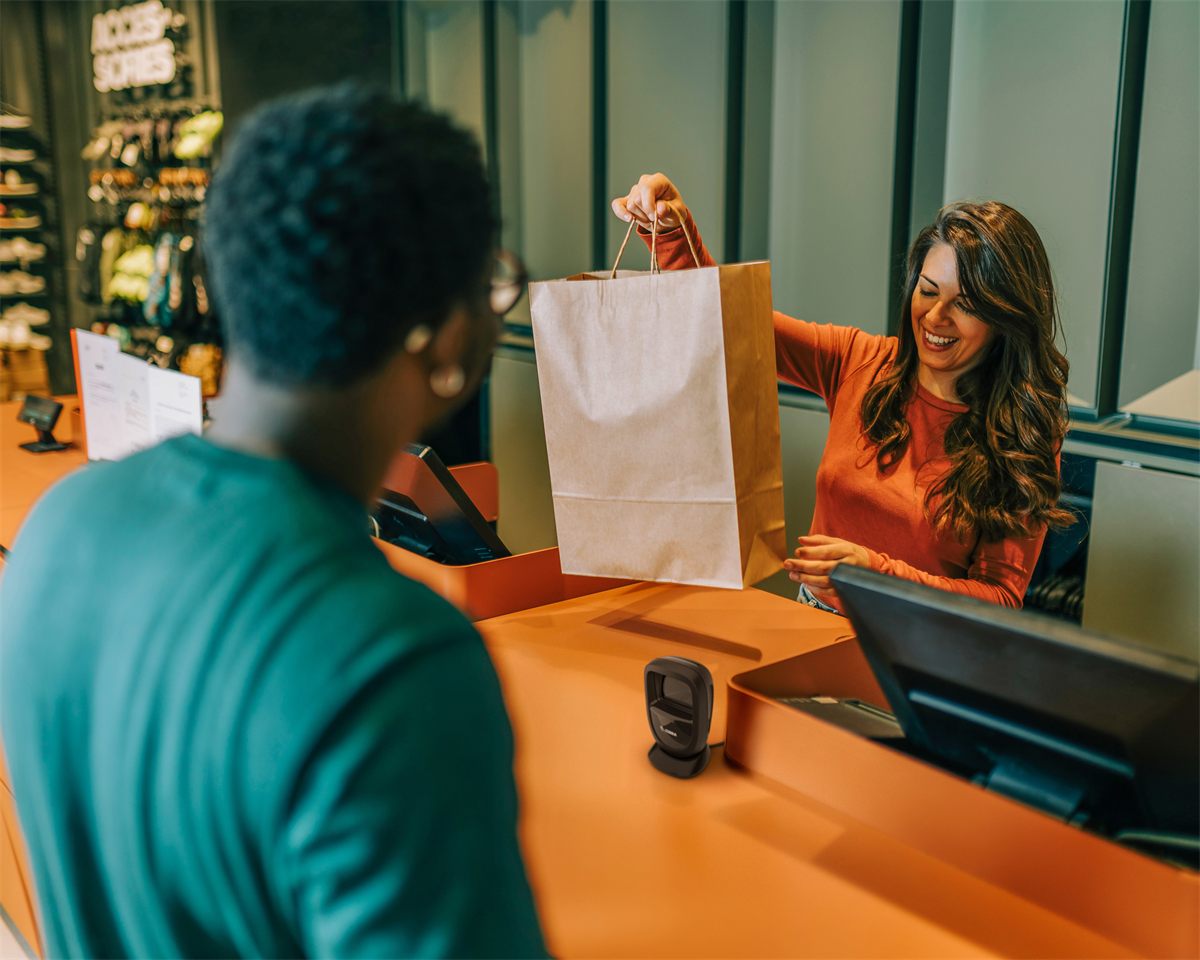 A shop assistant handing a customer their bag across a retail counter, with a DS9308 standing on the counter beside the till.